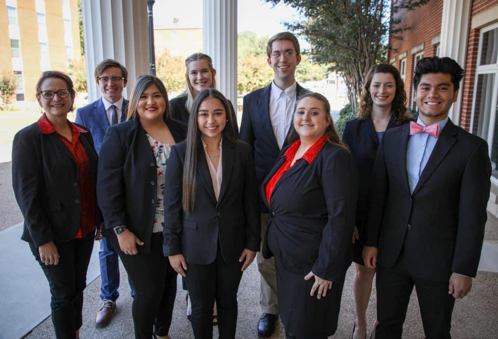 Group of eight well-dressed individuals smiling for a photo outside a building with columns at Howard Payne University. | HPU